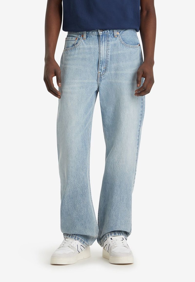 Homme portant un jean large bleu clair et des baskets blanches, debout devant un fond uni.