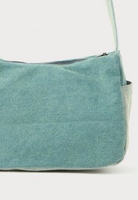 Teal denimaxelväska med dragkedjeslutning, mjuk textur och rund form. Har en justerbar rem och platt botten.