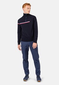 Pullover a maglia blu navy con colletto alto e zipper corta, presenta una striscia orizzontale rossa e bianca. Abbinato a pantaloni blu e scarpe scure.