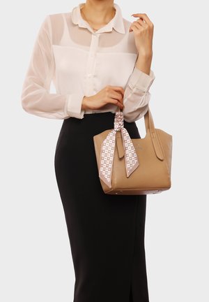 Borsa in pelle color tan con design strutturato, due manici e una sciarpa di seta con motivo legata attorno al manico. Donna che indossa una blusa bianca trasparente e una gonna nera.
