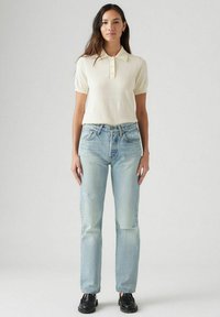 Polo crema a maniche corte con tre bottoni, abbinato a jeans azzurri chiari a gamba dritta e scarpe nere. Abbigliamento semplice e casual.
