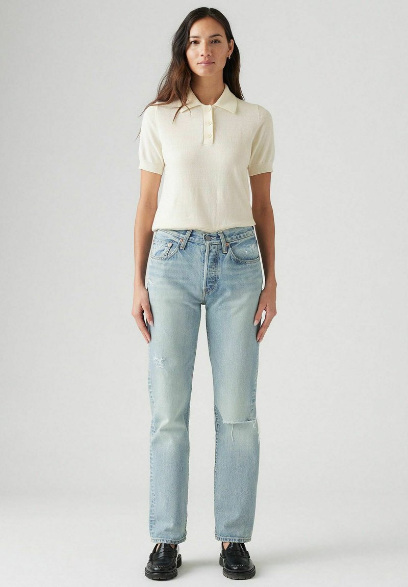 Polo crema a maniche corte con tre bottoni, abbinato a jeans azzurri chiari a gamba dritta e scarpe nere. Abbigliamento semplice e casual.