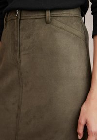 Gros plan sur une jupe en daim vert olive avec des détails de boutons et de coutures, portée avec un haut noir, montrant une partie de la main droite.