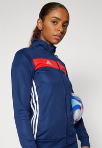 Giacca sportiva navy con dettagli rossi e bianchi, chiusura con zip e colletto alto. Il modello tiene un pallone da calcio multicolore sotto il braccio.