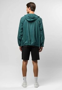Waterdichte groene jas met een trekkoordcapuchon, met een rechte snit en elastische manchetten. Draag het over zwarte shorts en witte sneakers.