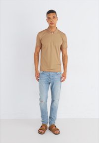 Camisa tipo polo beige de manga corta, con cuello de media cremallera, combinada con jeans rotos de color azul claro y sandalias marrones. Conjunto sencillo y casual.