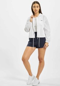 Valkoinen kevyt takki, jossa on joustavat hihansuut, vetoketjukiinnitys ja huppu, yhdistettynä tummansinisiin shortseihin ja valkoisiin urheilujalkineisiin.