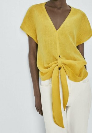 Dark-skinned person wearing a yellow short-sleeve V-neck top tied in a front knot with white high-waisted pants.