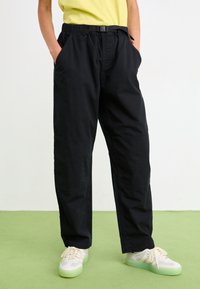 Schwarze, locker sitzende Hose mit elastischem Bund und Gürtel. Verfügt über zwei Seitentaschen und einen strukturierten Stoff. Getragen mit hellen Schuhen.