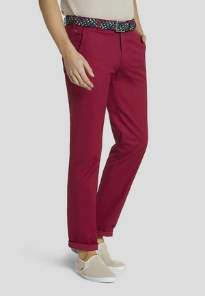 Mann trägt hochgekrempelte burgunderfarbene Hose, einen mehrfarbigen gewebten Gürtel, beige Slipper und ein hellbeiges, hineingestecktes Hemd.