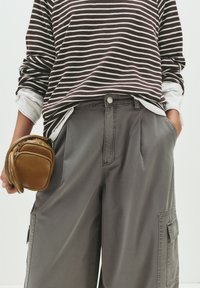 Pantaloni cargo grigi a gamba larga con tasche laterali, abbinati a una maglietta a maniche lunghe a righe marroni e bianche; piccola borsa marrone tenuta in mano.