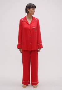 Červená saténová súprava pyžama obsahuje tričko s dlhými rukávmi, zapínanie na gombíky s vreckom a široké nohavice s bielymi lemami.