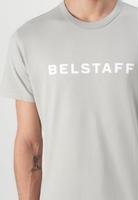 Γκρι βαμβακερό t-shirt με τολμηρό λευκό κείμενο "BELSTAFF" στο στήθος; κοντά μανίκια και στρογγυλή λαιμόκοψη. Λείος υφασμάτινος χαρακτήρας.