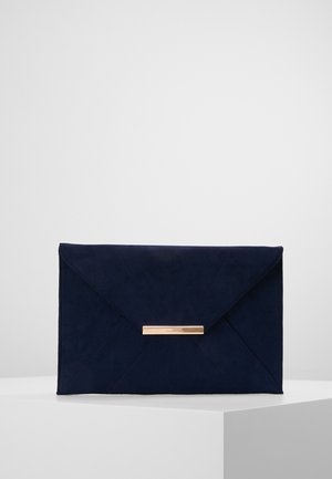 Pochette enveloppe en daim bleu marine avec rabat triangulaire et fermeture à barre métallique horizontale dorée, posée sur un piédestal blanc.