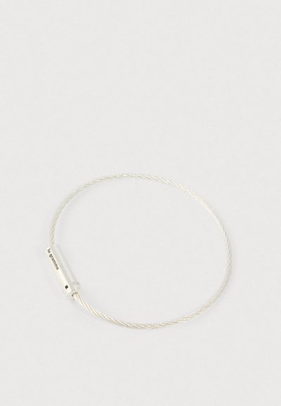 Srebrna spiralno uvijena narukvica s cilindričnom kopčom ugraviranom s "le gramme" na bijeloj pozadini.