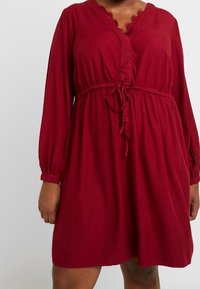 Abito rosso a maniche lunghe realizzato in tessuto morbido, con vita legata e rifinitura in pizzo lungo il scollo a V. Silhouette fluida con polsini elastici.