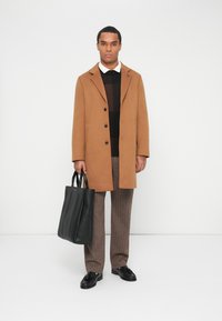 Uomo in soprabito marrone, maglione nero, pantaloni grigi e scarpe nere che tiene una grande borsa a tracolla in pelle nera su uno sfondo bianco.