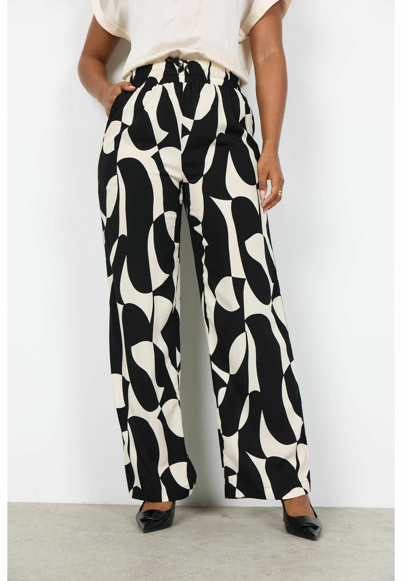 Kobieta ubrana w spodnie z wysokim stanem i szerokimi nogawkami w czarno-białe abstrakcyjne wzory, w połączeniu z czarnymi butami na spiczastym nosku oraz białą górą.