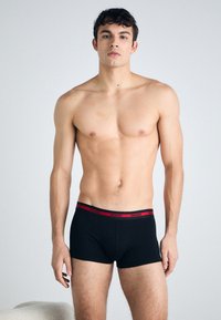 Sorte boksershorts med rødt livbånd som har "HUGO"-merke. Modellen har en muskuløs kroppsbygning og er uten skjorte mot en nøytral bakgrunn.