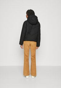 Doudoune noire avec une capuche, présentant une couture en chevrons. Associée à un pantalon en velours côtelé beige évasé et des chaussures blanches, vue de dos.