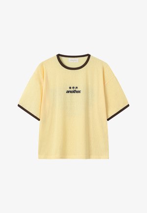 T-shirt jaune à manches courtes avec bordure marron foncé au niveau du col et des manches, avec le texte noir "another" et trois petites icônes au-dessus du centre de la poitrine.