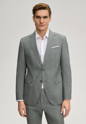 Jonge man die een lichtgrijs geruit nauwsluitend pak draagt met een wit overhemd, staand tegen een effen achtergrond, naar voren kijkend.