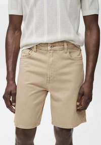 Beige denim shorts with a straight cut, featuring five-pocket styling and a metal button closure. Worn with a white knit top.