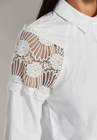 Witte katoenen blouse met ingewikkelde witte kantdetails op de schouder. Beschikt over een klassieke kraag en plooien voor extra textuur.