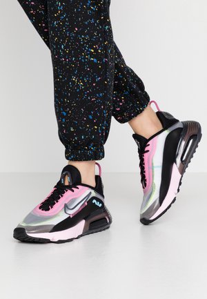 Persona che indossa pantaloni della tuta neri con macchie multicolori e sneaker nere, rosa e trasparenti che fa un passo avanti su un pavimento semplice.