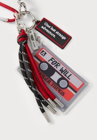 Atslēgu piekariņš ar kasetes dizainu sarkanos, melnos un baltos toņos. Ietver birku ar tekstu, pītu auklu un metāla aparatūras detaļas.