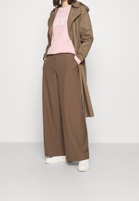 Pantalon large marron, sweat-shirt rose clair avec texte blanc, et un trench-coat marron. Des baskets blanches complètent la tenue.