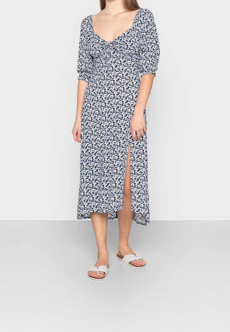 Midi šaty s květinovým potiskem v námořnické modři a bílé, se širokými rukávy, kulatým výstřihem a bočním rozparkem. Nosené se plochými sandály.