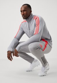 Grijs sportieve tracksuit met rode accenten, uitgerust met een hoge kraag en drie zwarte strepen. Witte voetbalschoenen maken de outfit compleet.