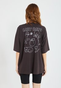Mørkebrun oversized t-skjorte med hvite linjete tegnede katter og teksten "LAZY DAYS" på baksiden. Mykt bomullsmateriale.