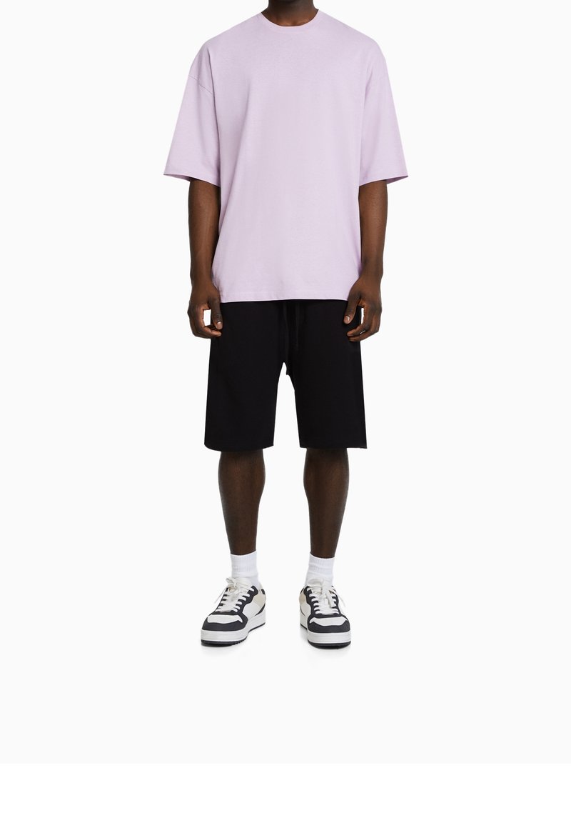 Homme portant un T-shirt ample violet clair, un short noir jusqu'aux genoux, des chaussettes blanches et des baskets noirs et blancs, debout devant un fond blanc.