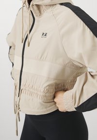 Beige vindjakke med sorte accenter, elastiske manchetter og snoretræk i taljen. Har et logo på brystet og en lynlåslukning foran.