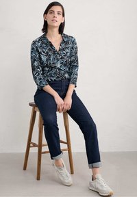 Blusa floreale a maniche lunghe in toni scuri con motivi blu e bianchi, abbinata a jeans di denim scuri e sneakers bianche, seduta su uno sgabello di legno.