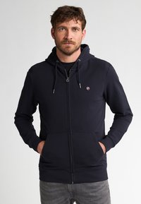 Marine blauwe rits-hoodie met een trekkoordcapuchon, voorzakken, geribbelde manchetten en een klein logo op de borst. Zachte stoftextuur.