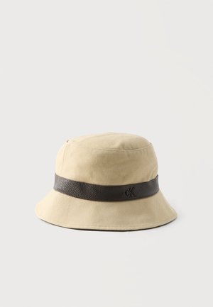 Chapeau bob beige avec un large bord et une bande texturée marron foncé ornée d'un petit logo "cK" en relief.