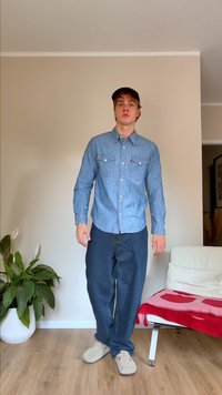 Šviesiai mėlynas džinsinis marškinėlis su mygtukais, turintis dvi krūtinės kišenes ir Levi's logotipą, derinamas su plačiomis tamsiomis kelnėmis ir smėlio spalvos atlenkiamaisiais bateliais.