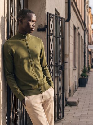 Homme en pull zippé vert et pantalon beige appuyé contre un mur sur un trottoir en ville avec des grilles en fer forgé derrière lui.