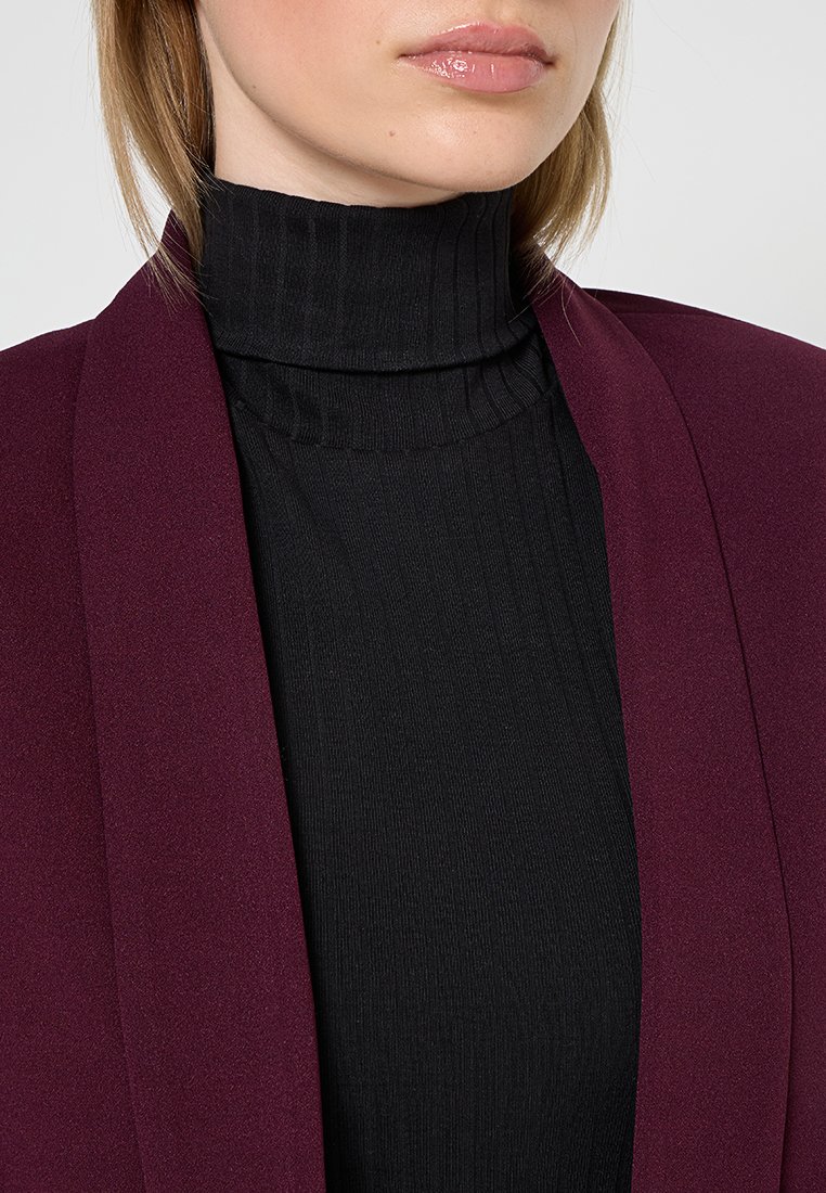 Blazer burdeos con un acabado suave, diseño de frente abierto, superpuesto sobre un cuello de tortuga negro con textura acanalada. Enfoque en la textura y el contraste de colores.