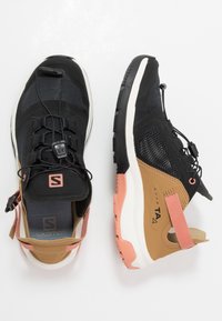 Le scarpe sportive nere e marroni presentano una tomaia in rete, lacci regolabili, una cinghia di accentuazione posteriore e una suola rosa con un motivo testurizzato.