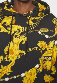 Zwarte hoodie met sierlijke gouden bloemen- en kettingpatronen, met een merklabel en trekkoorden. Zachte stof met levendige details.