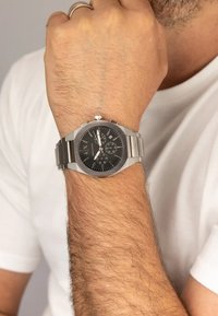 Orologio in acciaio inox con quadrante nero, dettagli in tonalità argento, funzioni cronografiche e bracciale a maglie, indossato su un polso con peli leggeri.