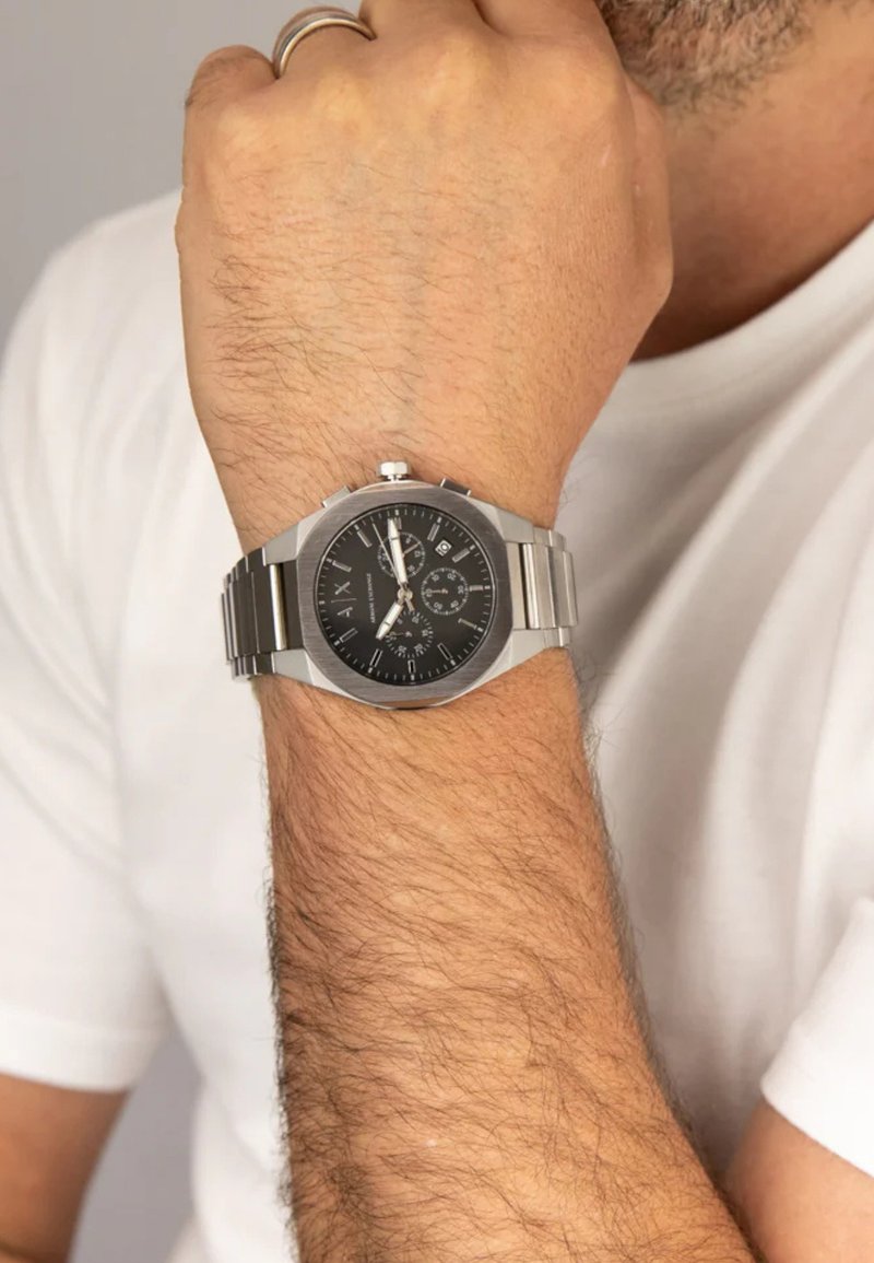 Orologio in acciaio inox con quadrante nero, dettagli in tonalità argento, funzioni cronografiche e bracciale a maglie, indossato su un polso con peli leggeri.