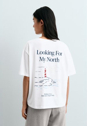 Vrouw met lang donker haar die een wit T-shirt draagt met de tekst "Looking For My North" en een vuurtorenillustratie op de rug.