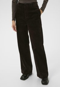Pantalons larges en velours côtelé marron avec des c�ôtes verticales. Ils présentent une taille haute et des poches latérales, associés à des chaussures noires.