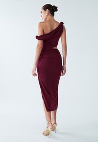 Burgunderrotes Midikleid aus elastischem Stoff, mit Off-Shoulder-Design, seitlichem Ausschnitt und hohem Schlitz. Goldfarbene Riemchenabsätze sichtbar.