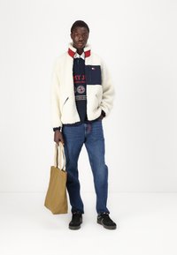 Giacca in pile color crema con colletto rosso e dettagli blu navy, abbinata a un maglione blu navy, jeans blu e una borsa a tracolla color sabbia. Sneakers nere completano il look.